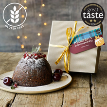 Load image into Gallery viewer, A Christmas pudding on a plate with cranberries and a sprig of rosemary, shown beside a wrapped gift box. The pudding has a Gluten Free badge overlay, and the gift box has a 'Great Taste 2026' award badge.
